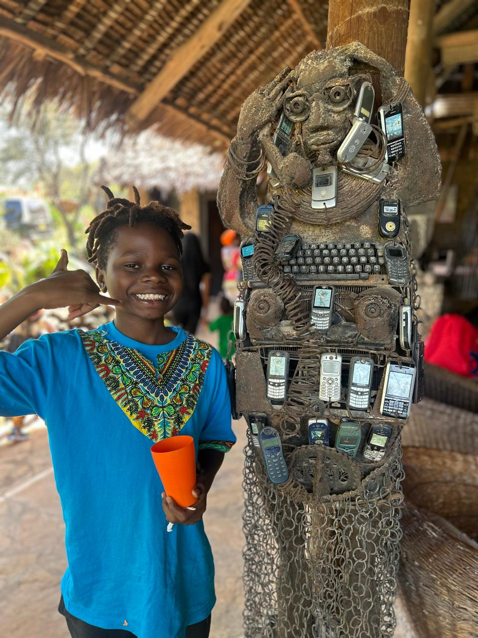A child in a blue dashiki-style shirt making a call me hand gesture next to a sculpture made of old phones and electronics.